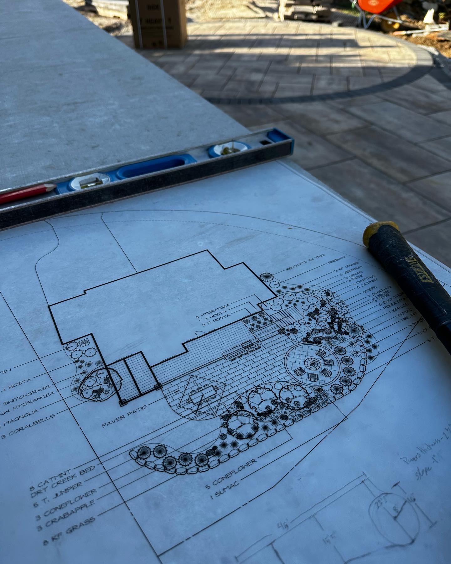 Blueprint of a paver patio design with landscaping details, including plant names and layout plans, set on a construction site with tools and materials visible.