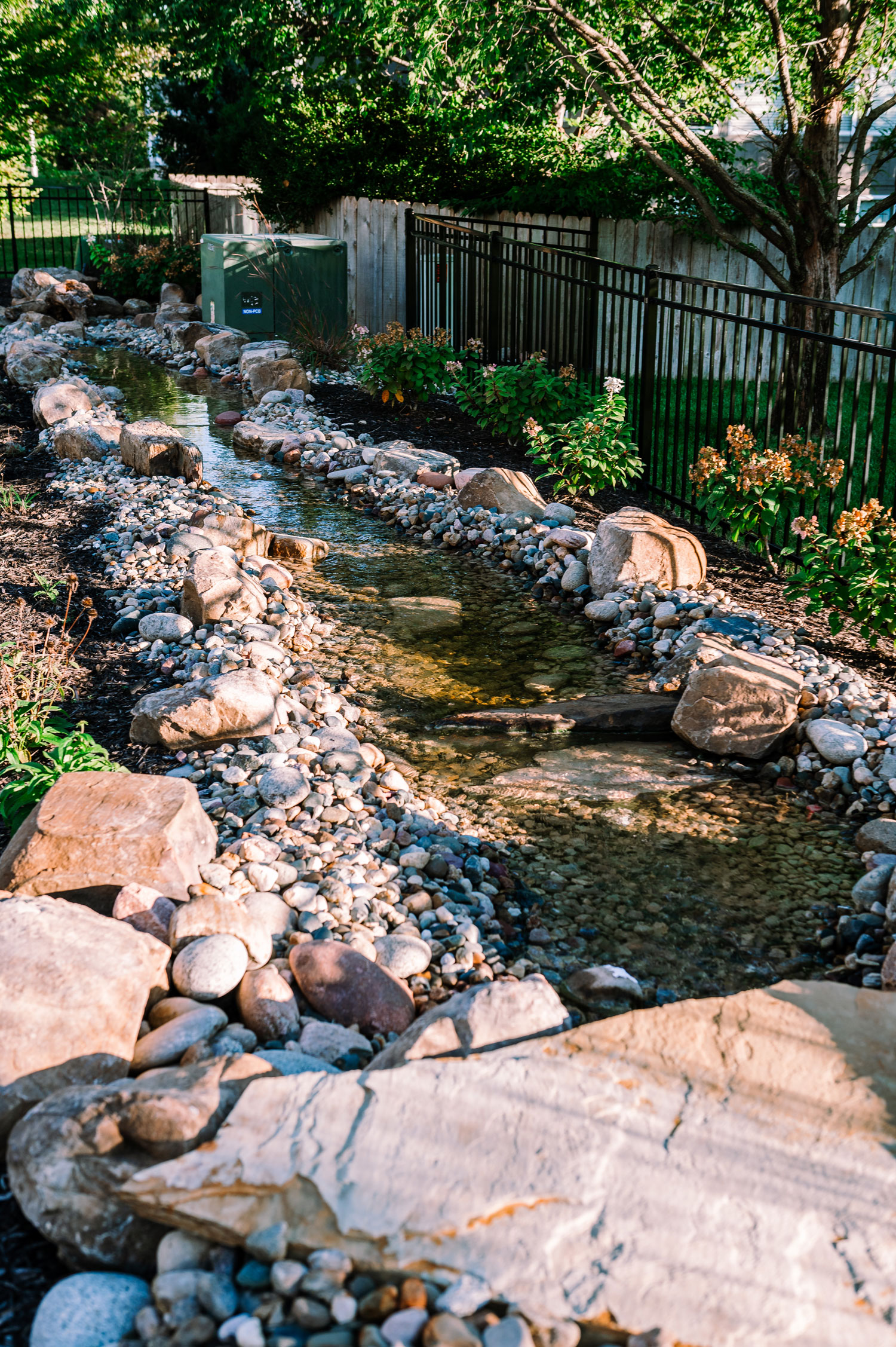 Stream with natural stones and pebbles, bordered by lush greenery and a black fence, creating a tranquil water feature for backyard serenity.