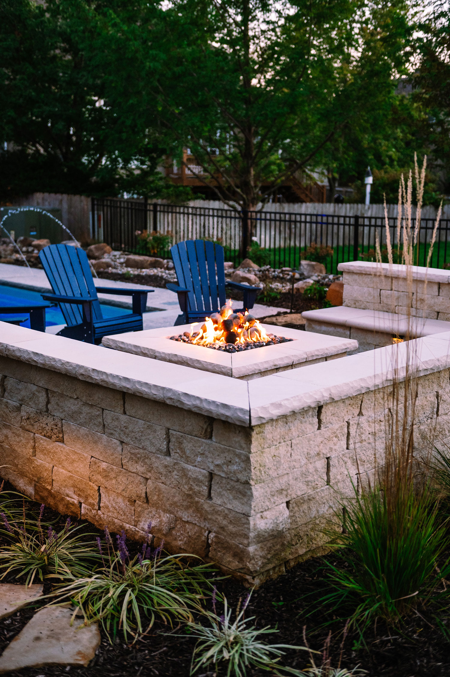 Outdoor fire pit with flames, surrounded by stone seating wall and blue Adirondack chairs, in a landscaped backyard setting with greenery and a pool.