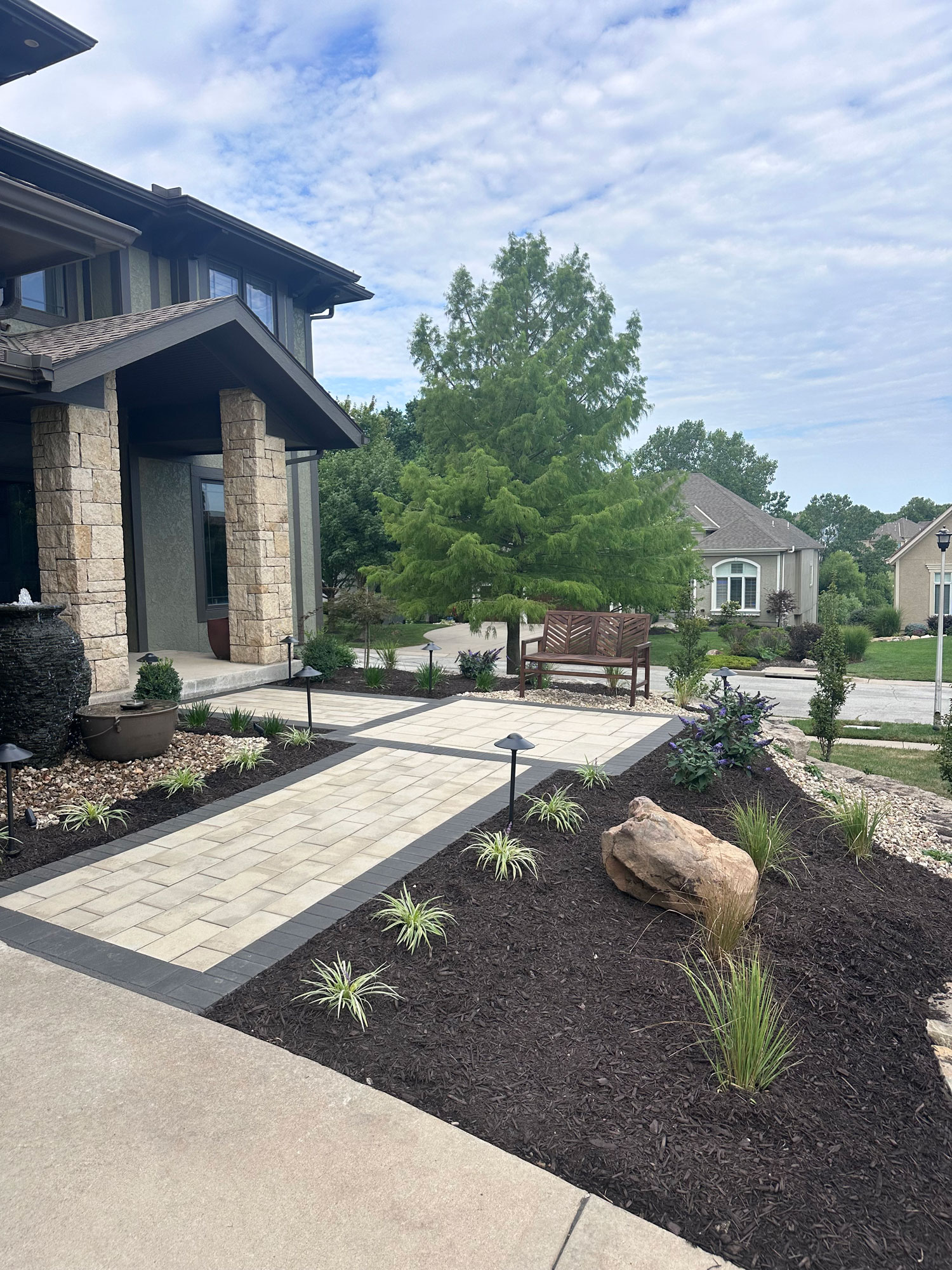 Custom paver patio installation featuring a stone walkway, decorative landscaping with plants and mulch, and a wooden bench in a residential outdoor setting.