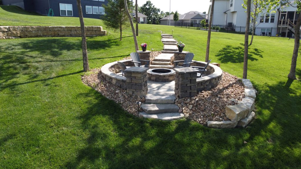 Outdoor living space featuring a circular stone fire pit surrounded by seating, landscaped with grass and decorative stones, highlighting hardscape design and functionality.