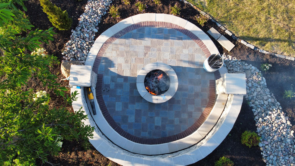 Circular stone patio with a fire pit, surrounded by landscaped greenery and decorative pebbles, showcasing outdoor living space design by Atlas Landscape.
