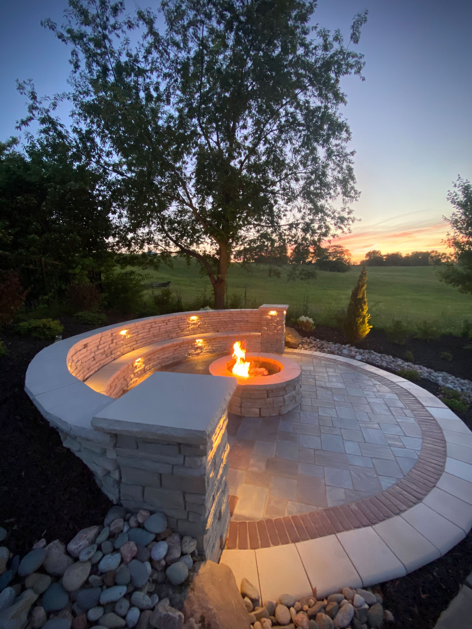 Outdoor fire pit installation with stone seating and illuminated features, surrounded by landscaped greenery and a sunset sky, showcasing Atlas Landscape's hardscaping expertise.