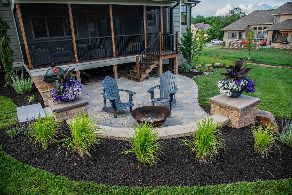 Outdoor living space featuring stone patio, fire pit, and Adirondack chairs surrounded by lush landscaping and flower planters, enhancing Kansas City home aesthetics.