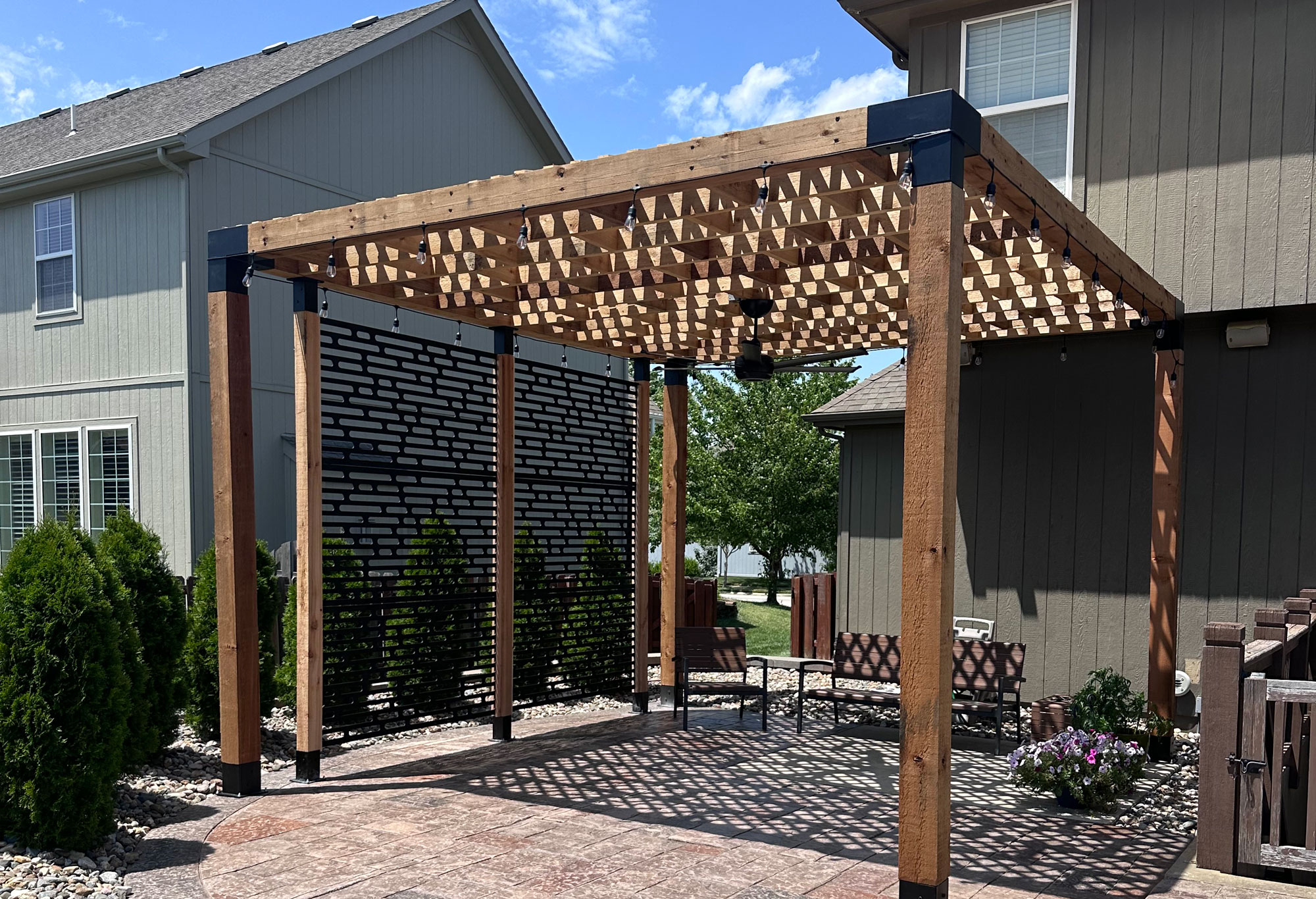 Custom wood pergola with decorative lattice design, surrounded by landscaped greenery and seating area, enhancing outdoor living space in a residential setting.